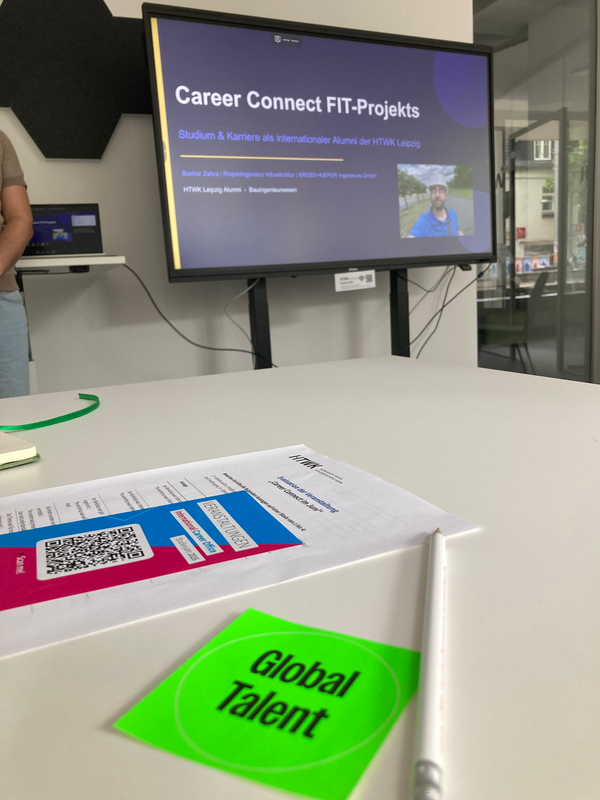 Zettel und Bleistift auf einem Arbeitstisch, im Hintergrund eine Präsentation
