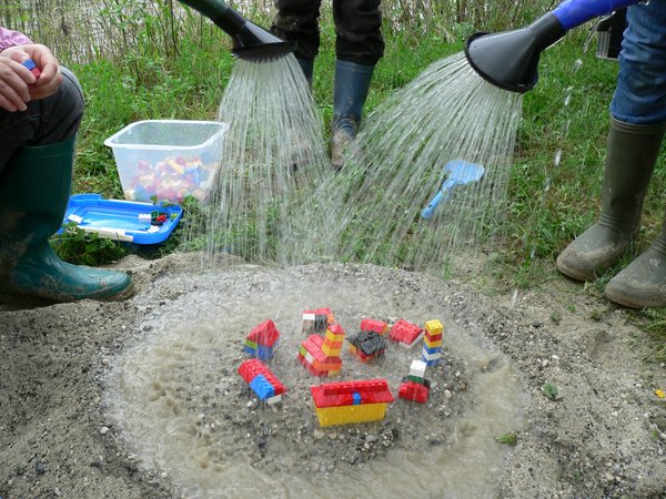 mehrere Personen gießen auf Spielfiguren in einem Sanskasten