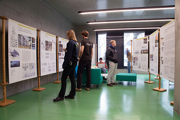 Im Foyer in der Hochschulbibliothek stehen Ausstellungstafeln, Personen besichtigen diese