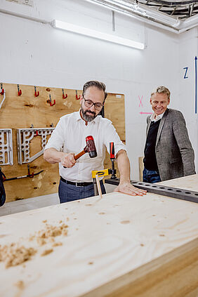 Sebastian Gemkow hämmert eigenhändig einen Holzdübel in eine Holzwandkonstruktion, neben ihm steht Prof. Alexander Stahr
