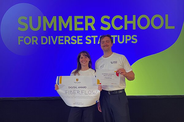 Lena und Ludwig von FiberFlow mit dem Gewinnercheck. Im Hintergrund große Leinwand mit der Aufschrift "Summer School for diverse Startups"