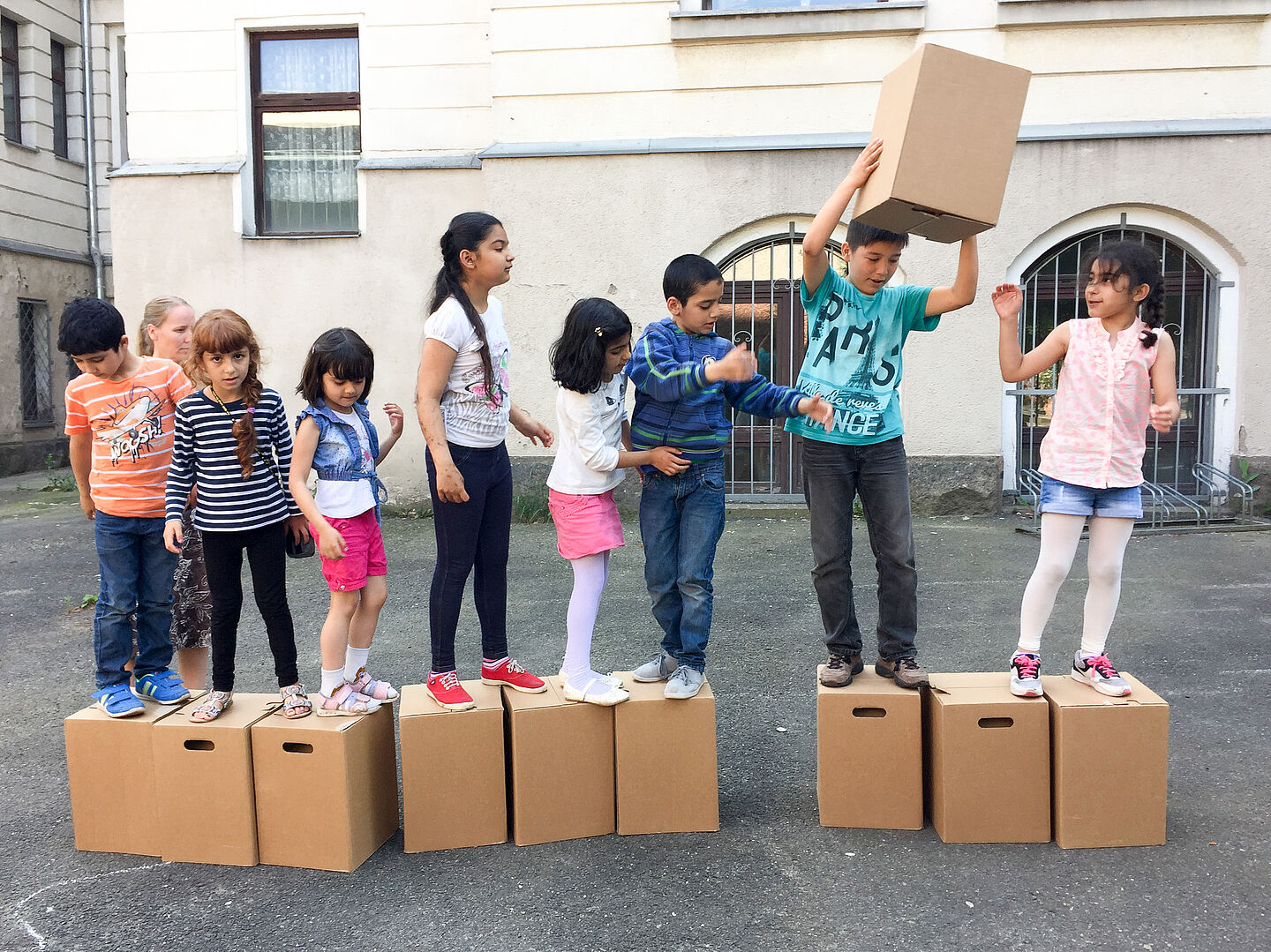 Mehrere Kinder stehen in einer Reihe auf jeweils einem Pappkarton. Eines der Kinder gibt einen Pappkarton durch die Reihe nach vorne.