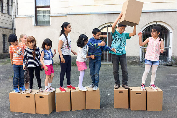 Mehrere Kinder stehen in einer Reihe auf jeweils einem Pappkarton. Eines der Kinder gibt einen Pappkarton durch die Reihe nach vorne.