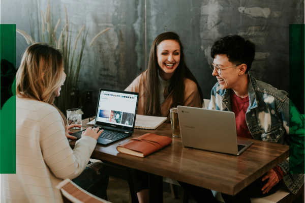 Drei Frauen sitzen zusammen am Tisch und reden miteinander. Vor ihnen stehen Laptops, Bücher und Getränke.