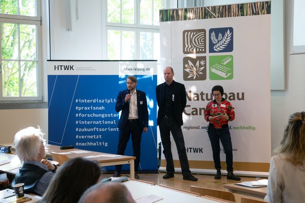 3 Personen stehen vor großen Roll-ups und sprechen in ein Mikrofon