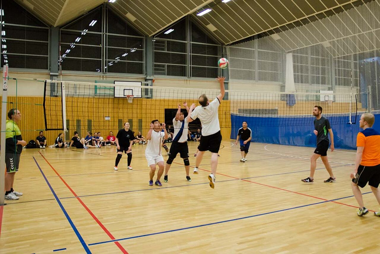 Menschen spielen in einer Sporthalle Volleyball. Einer von ihnen sprint in die Luft, um den Ball anzunehmen.