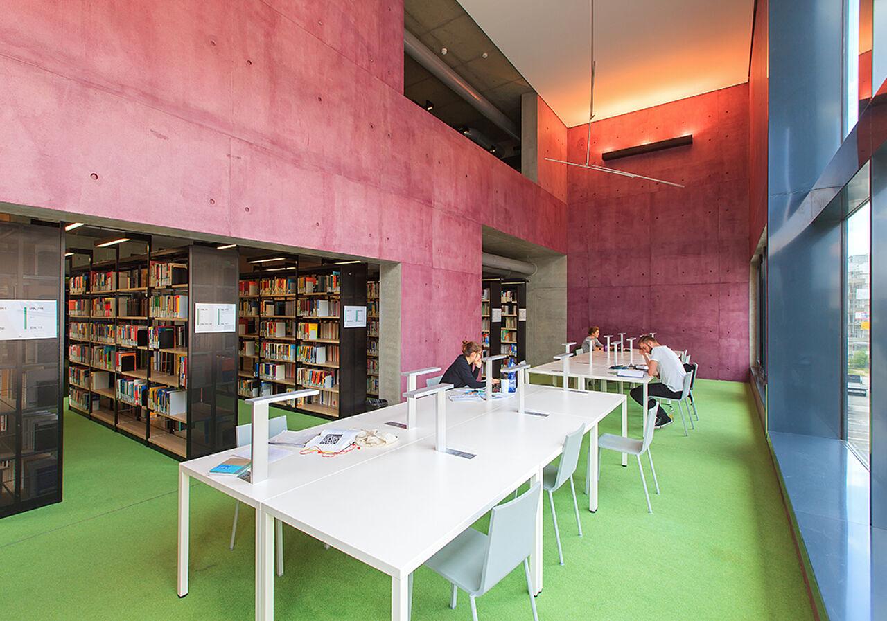 Das Bild ist in der Bibliothek der HTWK Leipzig aufgenommen worden. Es ist ein Lernbereich mit weißen Tischen zu sehen. Rechter Hand ist eine Fensterfront und linker Hand sind Bücherregale.
