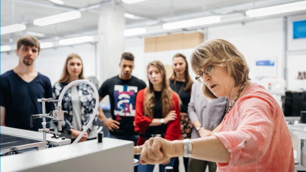 Eine Professorin erklärt Studierenden eine Druckmaschine. Dazu steht dort geschrieben: "Gestalten Sie die Zukunft mit!"